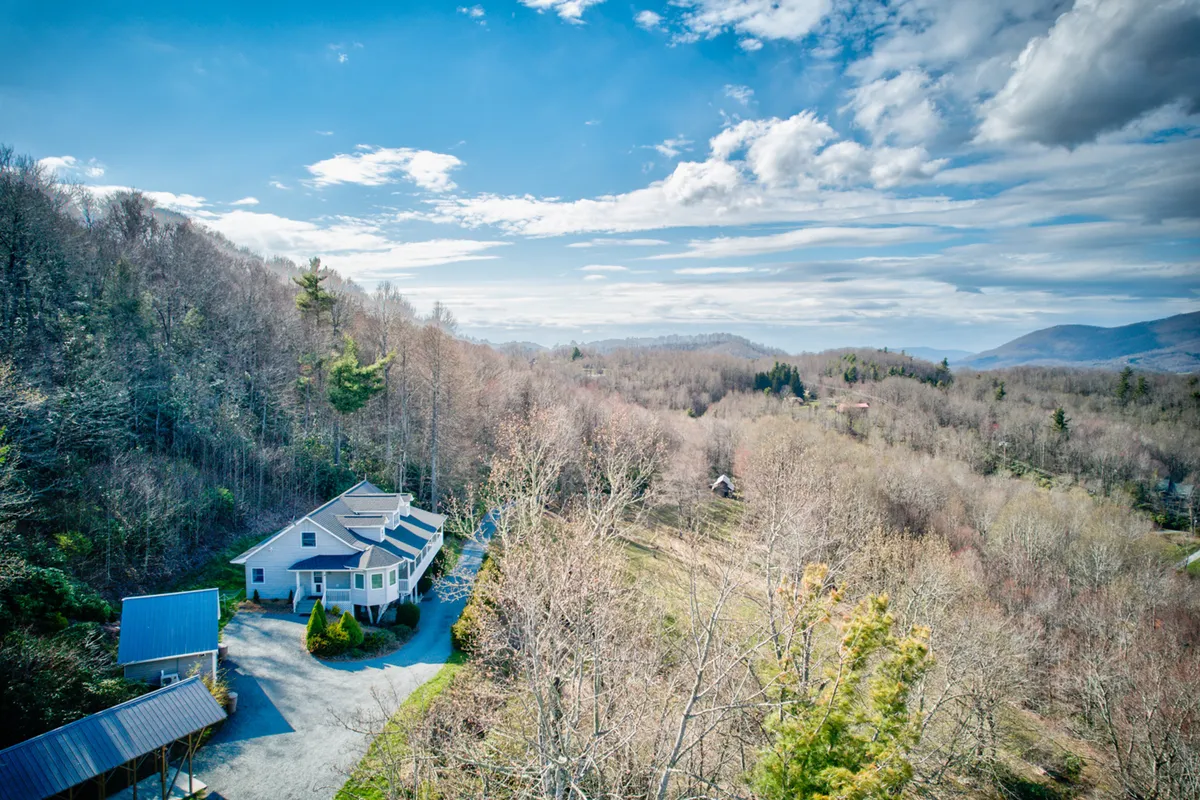 A sweeping exterior view of the house nestled amidst mature trees, with the driveway leading in and expansive mountain vistas in the background. The property’s open lawn and the forested surroundings convey a sense of privacy and immersion in nature.