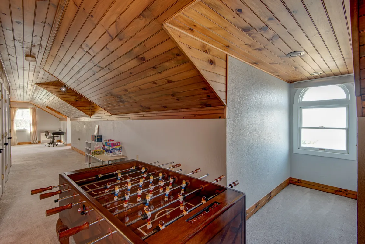 A bright, airy game room featuring a foosball table, large windows letting in natural light, and a wooden ceiling that adds warmth to the space. The open layout suggests an inviting atmosphere for friendly competition and socializing.