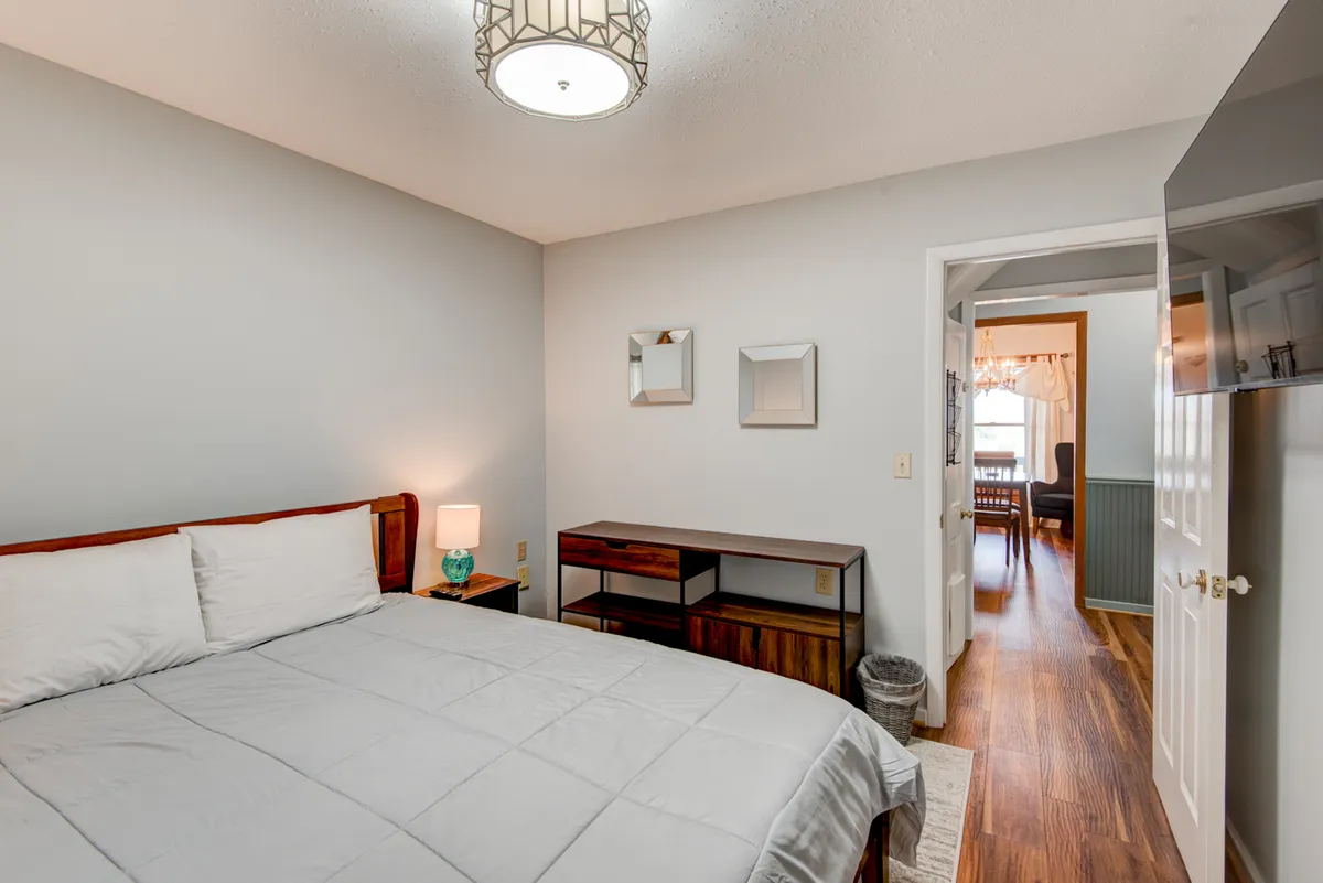A cozy bedroom scene with white bedding, a wooden nightstand, a decorative lamp, and tasteful wall art. The room feels intimate, with an open door hinting at additional spaces beyond.
