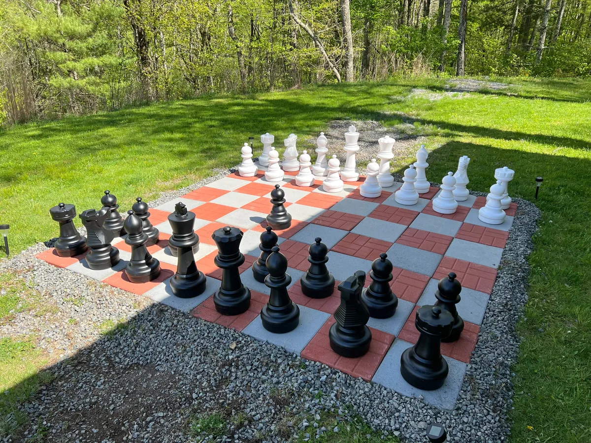 A vibrant grassy lawn with a life-size outdoor chess set, bordered by lush trees, creating a playful and interactive spot for both kids and adults. The open space encourages group activities and friendly competition.