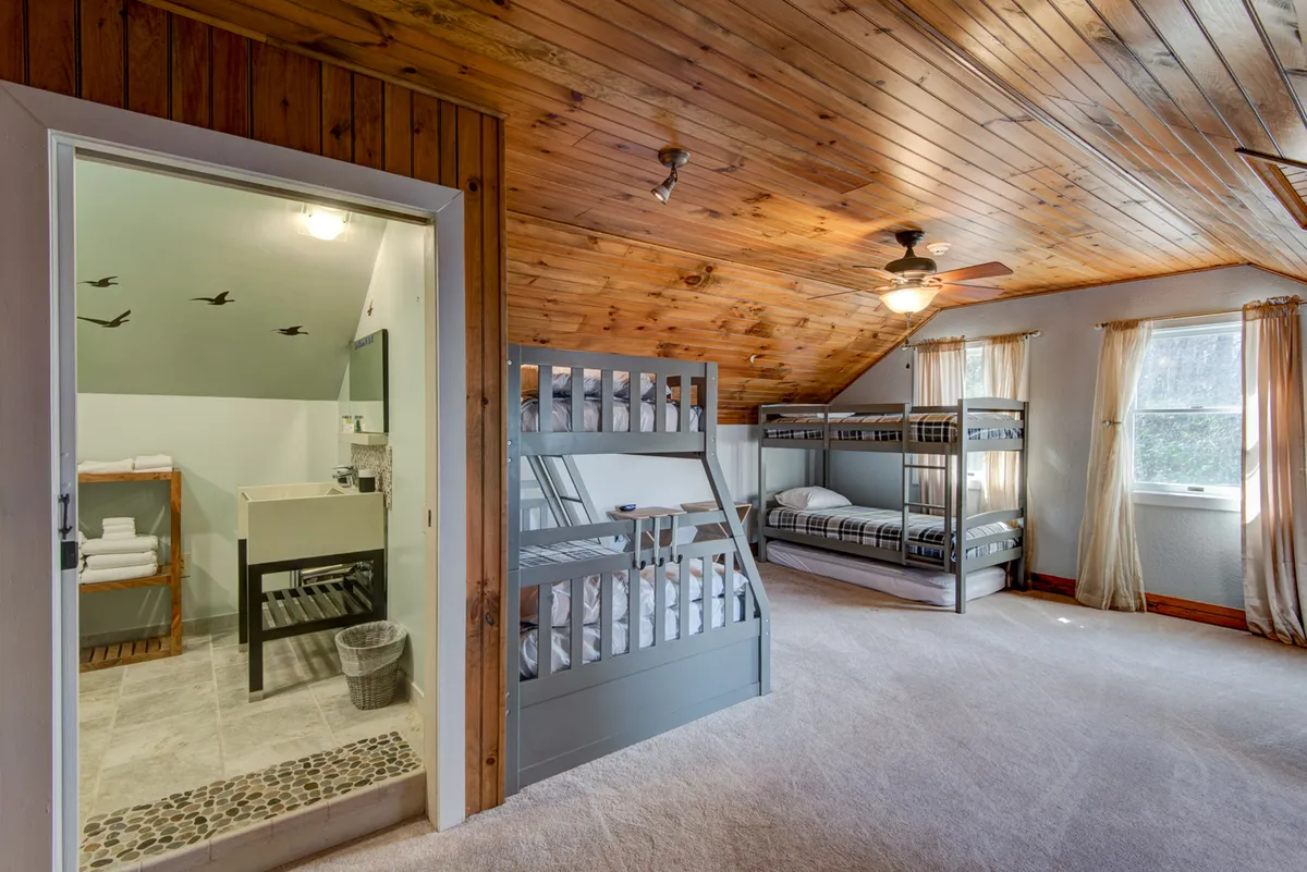 An inviting upstairs bunk area showcasing multiple bunk beds, a wooden ceiling, and an entrance to a bathroom. The space is bright, practical, and designed for flexibility, making it perfect for families or groups of friends.