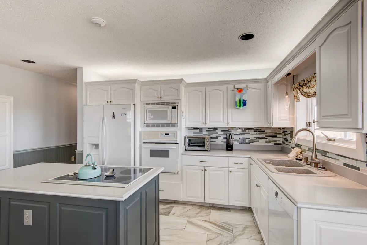 A bright, modern kitchen with a central island topped with a teal kettle, surrounded by sleek white cabinetry and state-of-the-art appliances. Decorative glass items on the window ledge and ample surface space make the kitchen functional and inviting, ideal for preparing meals or sharing casual conversations while cooking.