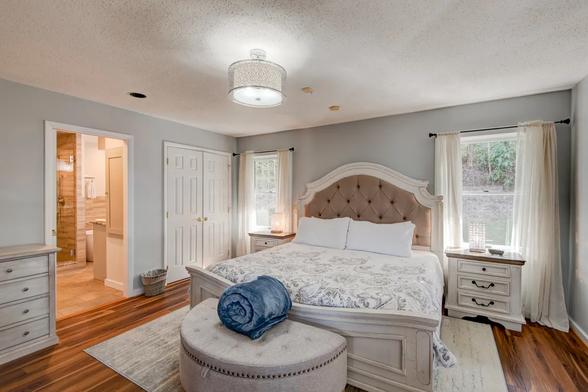 This image features a serene master bedroom with a king-sized bed, a stylish ottoman, and large windows letting in ample natural light. The decor is calm and inviting, perfectly suited for restful nights and peaceful mornings.